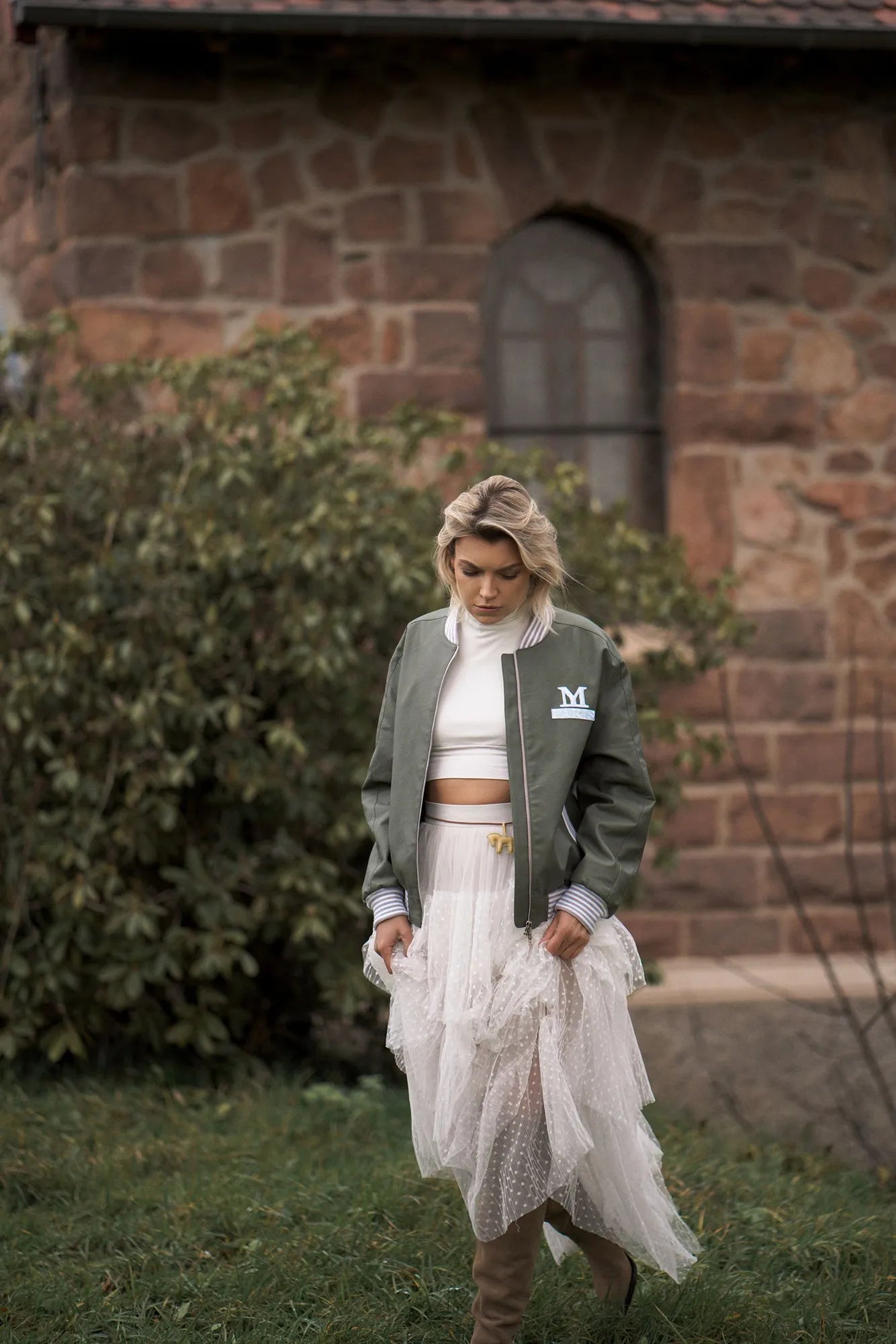 A woman wearing a white dress and green bomber jacket strolls in front of a stone building, reflecting 90s Paris fashion.