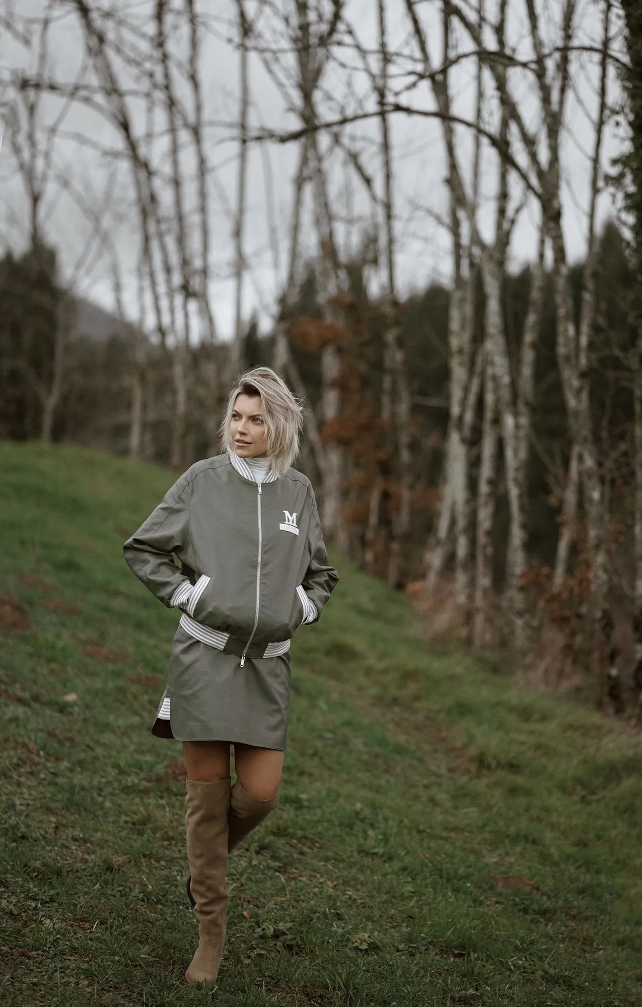 A woman in a green raincoat and boots walks on a grassy hill, showcasing a 90s style bomber jacket from Mimaussy Paris.