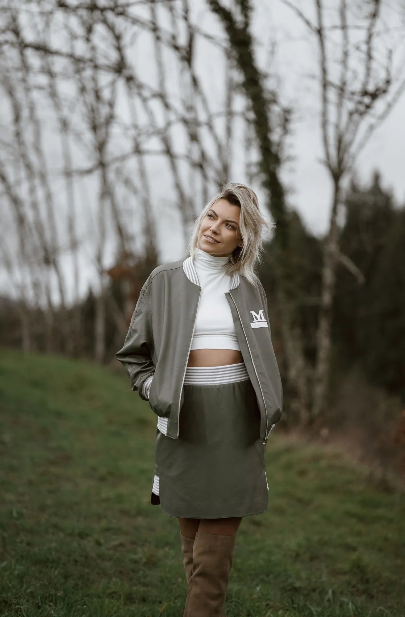 A woman wearing a green 90s-style Mimaussy Paris bomber jacket and skirt stands in a grassy field.