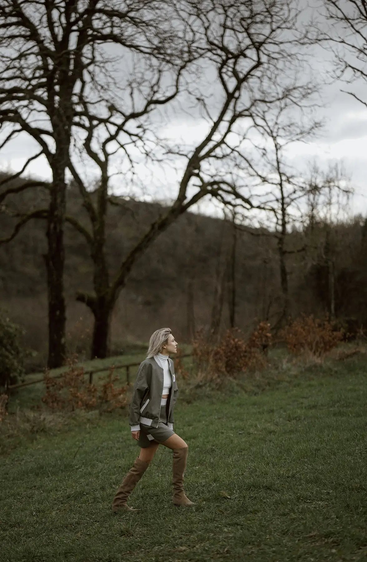 A woman in a green jacket and boots runs through a field, showcasing a Mimaussy Paris 90s style skirt.