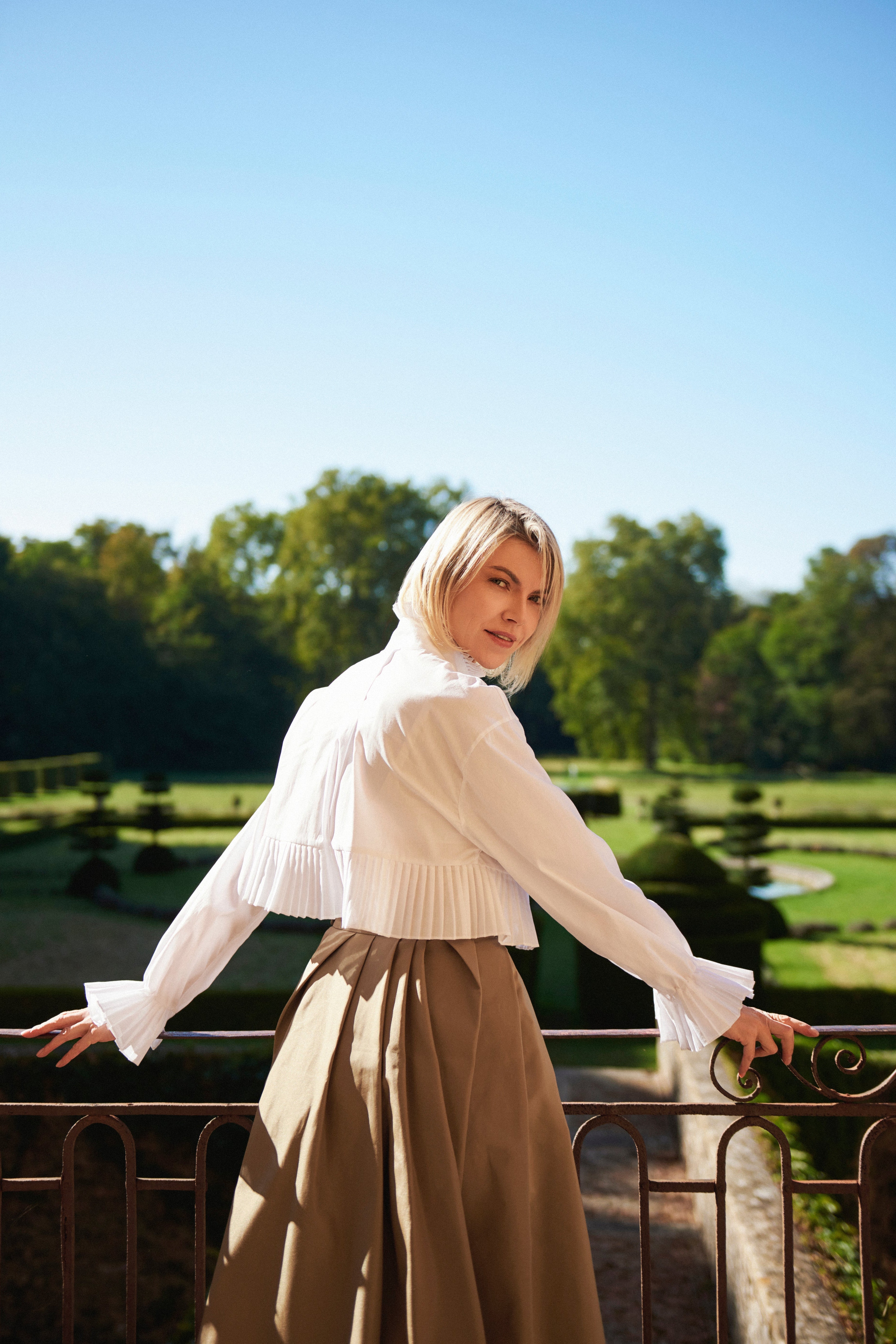 Woman wearing Mimaussy Paris Mademoiselle Plissé cropped blouse, white blouse and beige skirt, standing on a balcony with a scenic background
