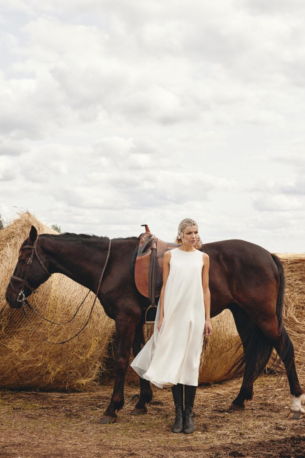 Woman in a white dress standing next to a horse in a field with hay bales.