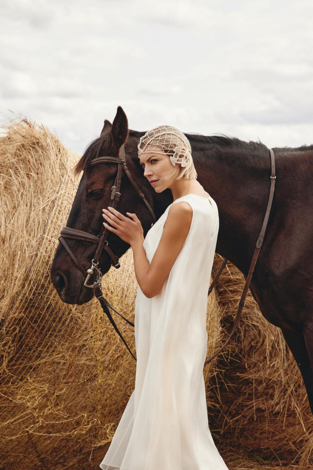 Woman in a white dress standing next to a horse in a field with hay bales.