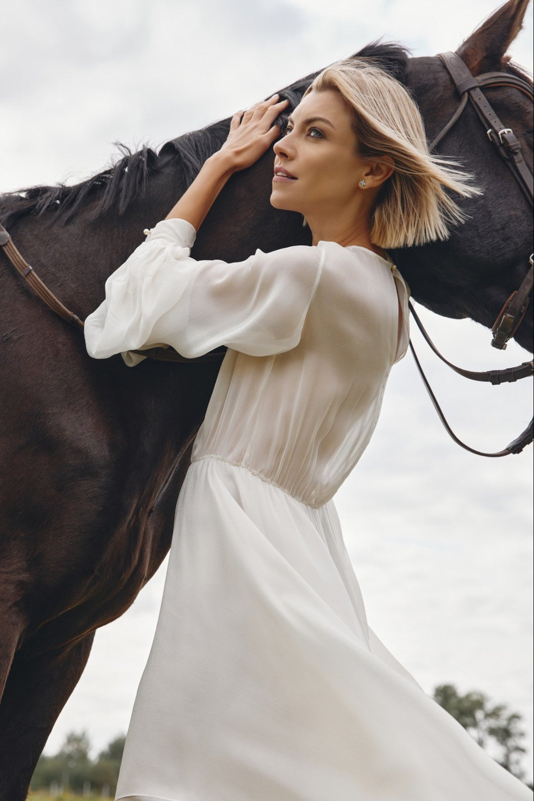 Woman in a white dress standing next to a horse with a cloudy sky background