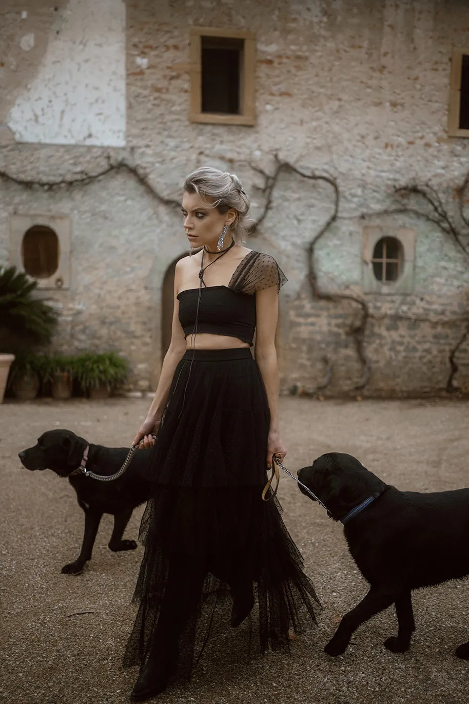 A woman in a black Souflé Skirt from Mimaussy Paris stands with two black dogs beside her.