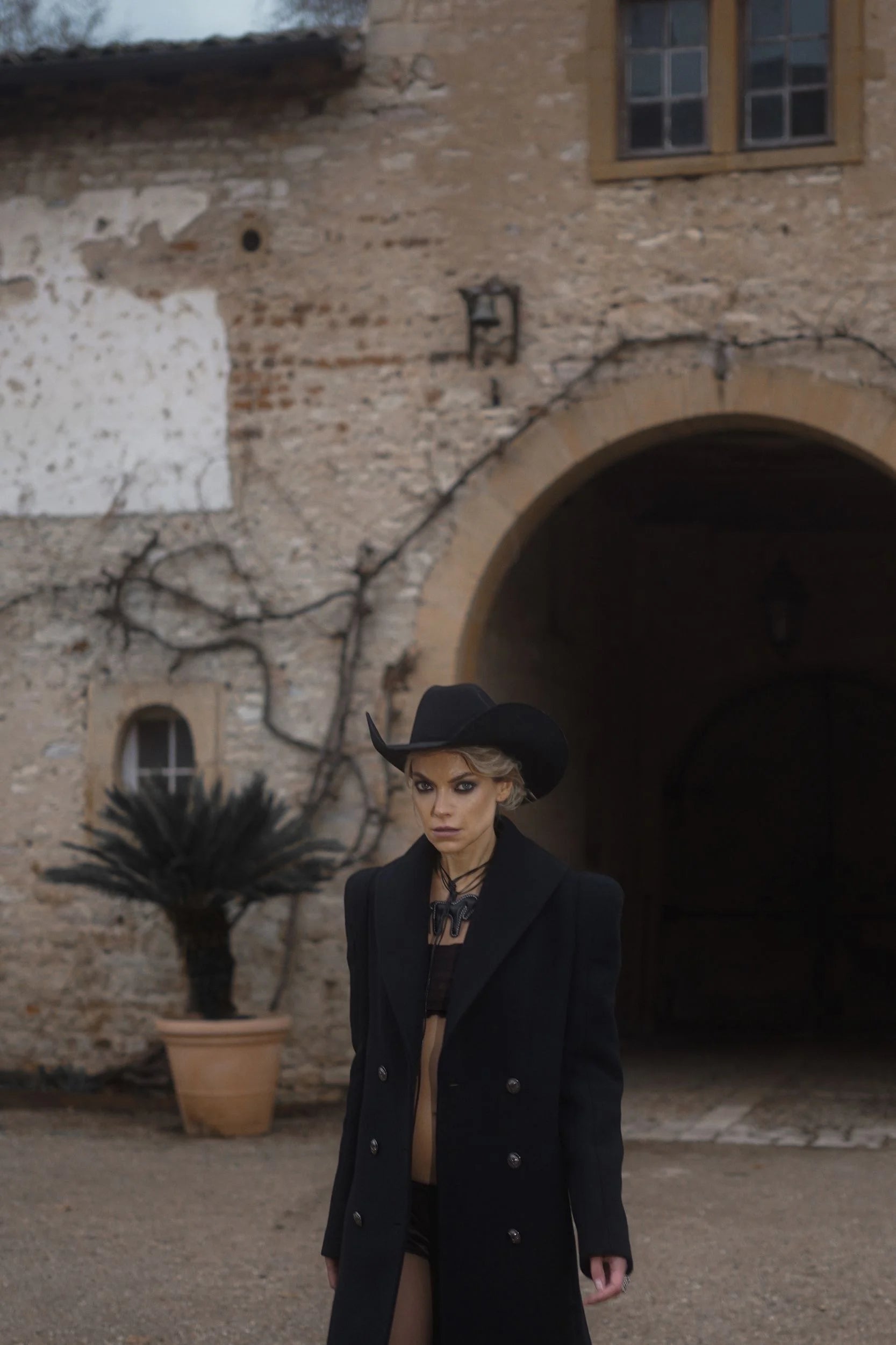 A woman wearing a black The Griffon coat and hat from the luxury brand Mimaussy Paris stands in front of a stone building, exuding elegance and style