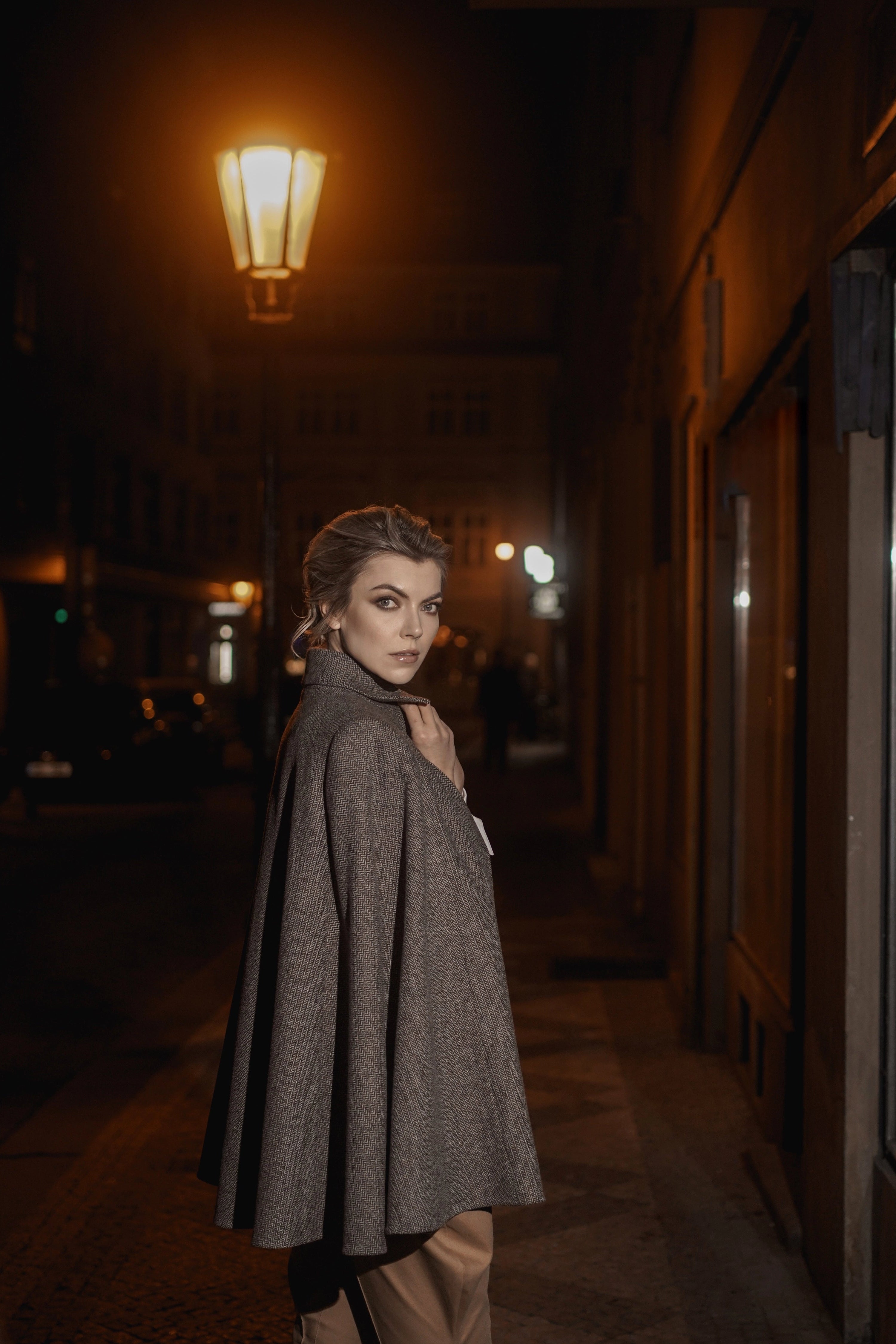 Woman wearing Cape “Brume” from Mimaussy Paris, gray coat standing on dark street at night illuminated by streetlight outdoors