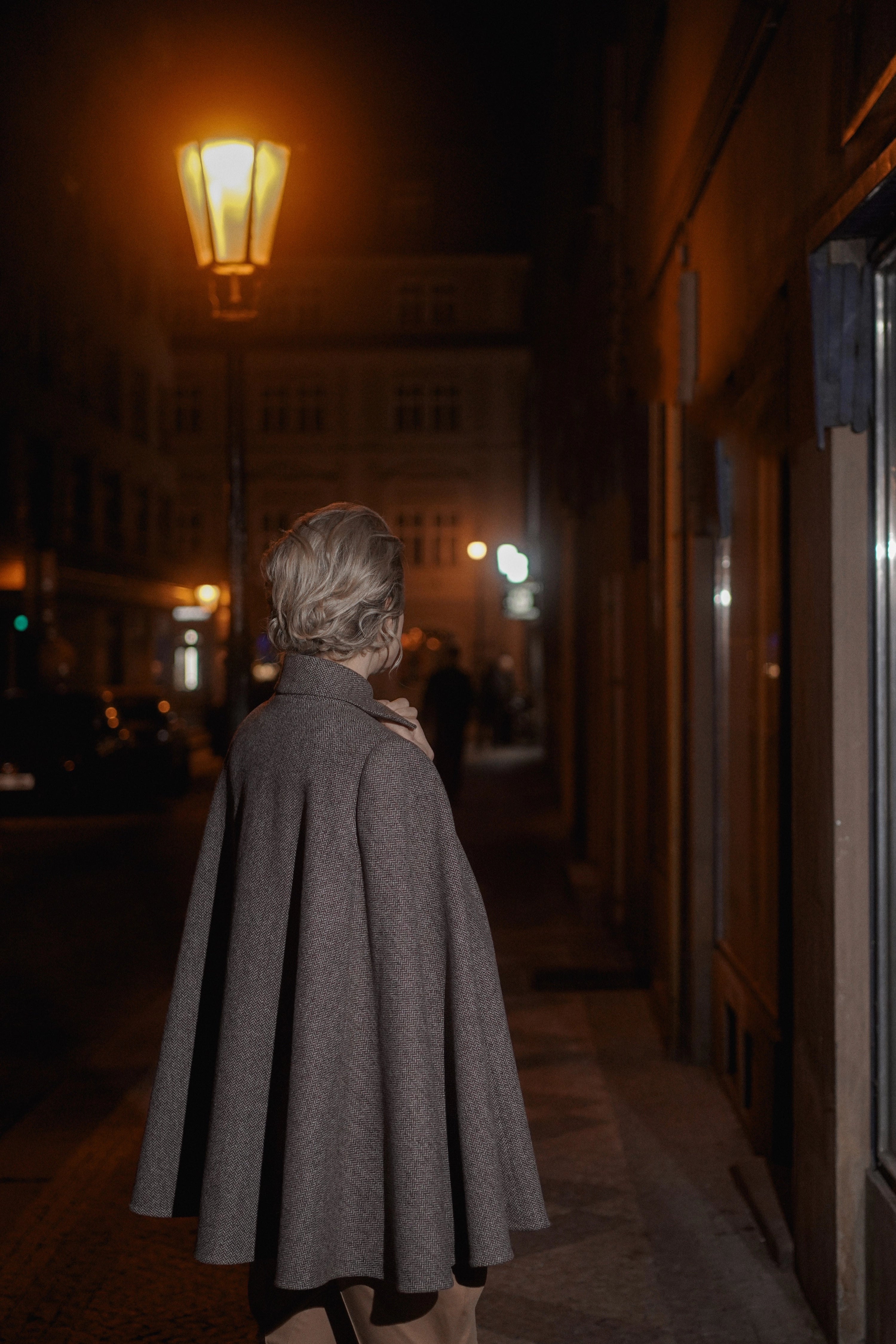 Woman wearing Cape “Brume” from Mimaussy Paris, gray coat while walking down street at night with warm light source outdoors