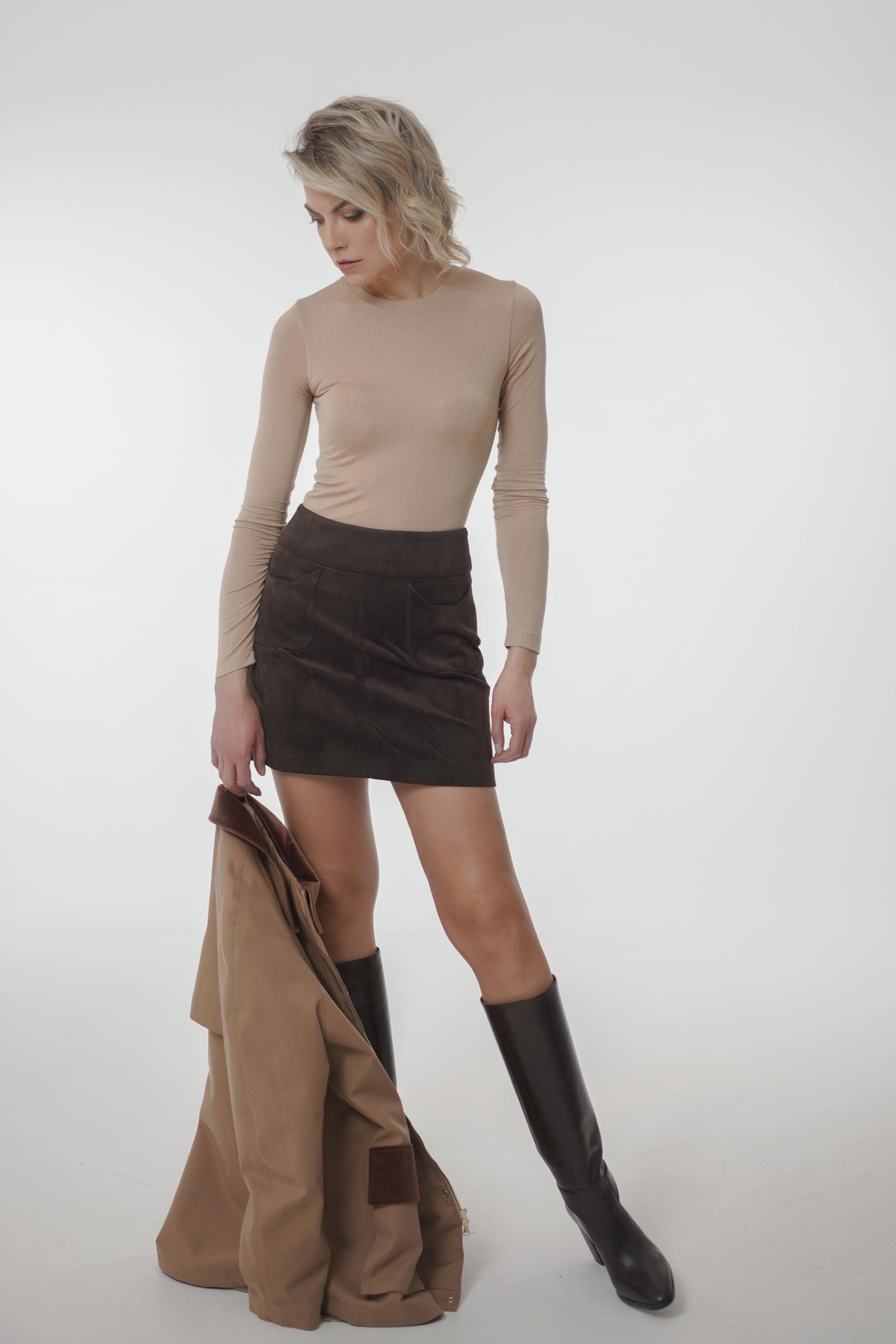 Woman wearing Catty Velour Skirt from Mimaussy Paris, brown skirt with black knee-high boots holding brown jacket on white background