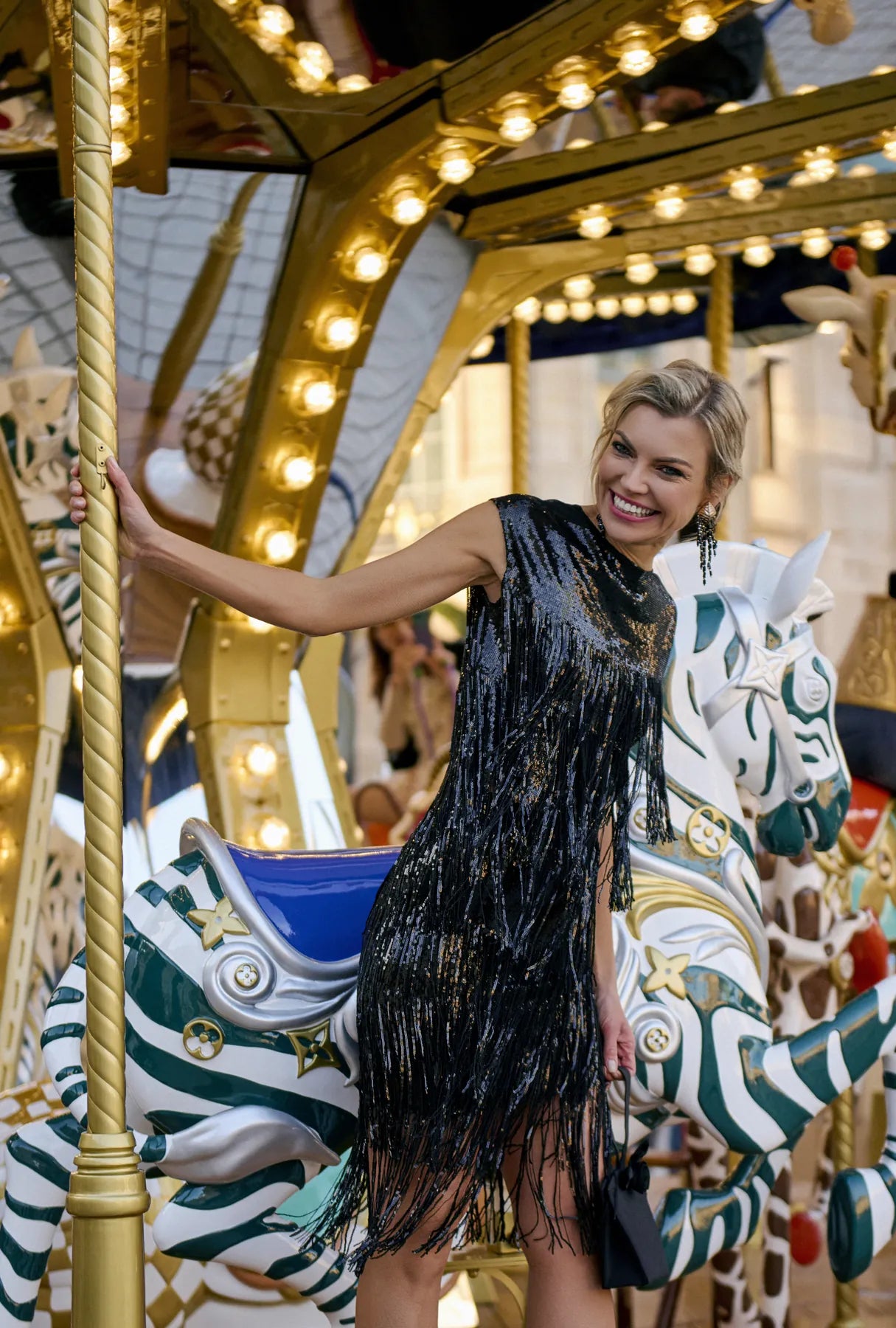 A woman in a stylish black dress poses on a carousel, highlighting the Charleston Glow Top&Skirt Set from Mimaussy Paris.