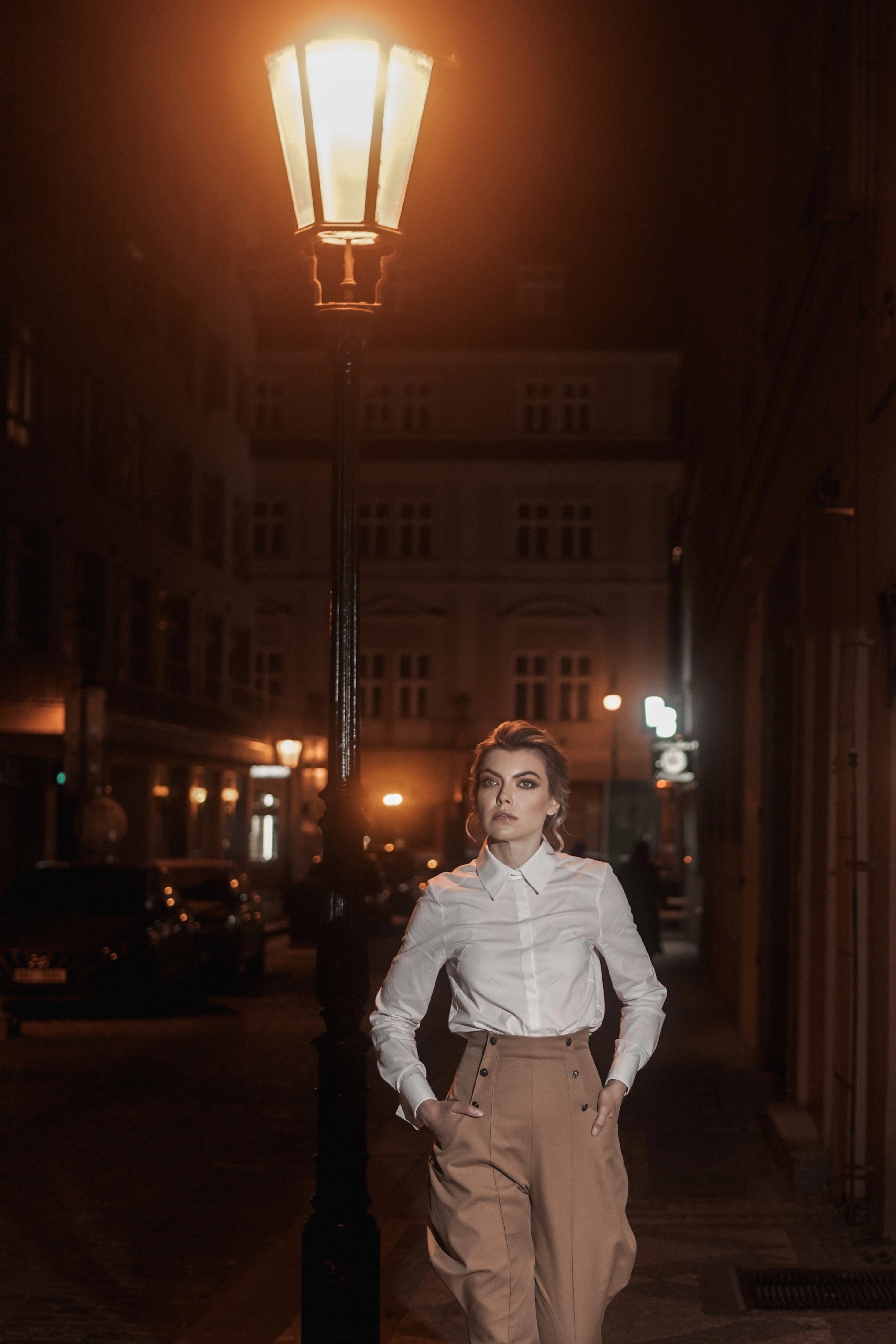 Woman wearing Cotton shirt “Discret” in white from Mimaussy Paris, standing on street at night with vintage street lamp and building in background outdoors