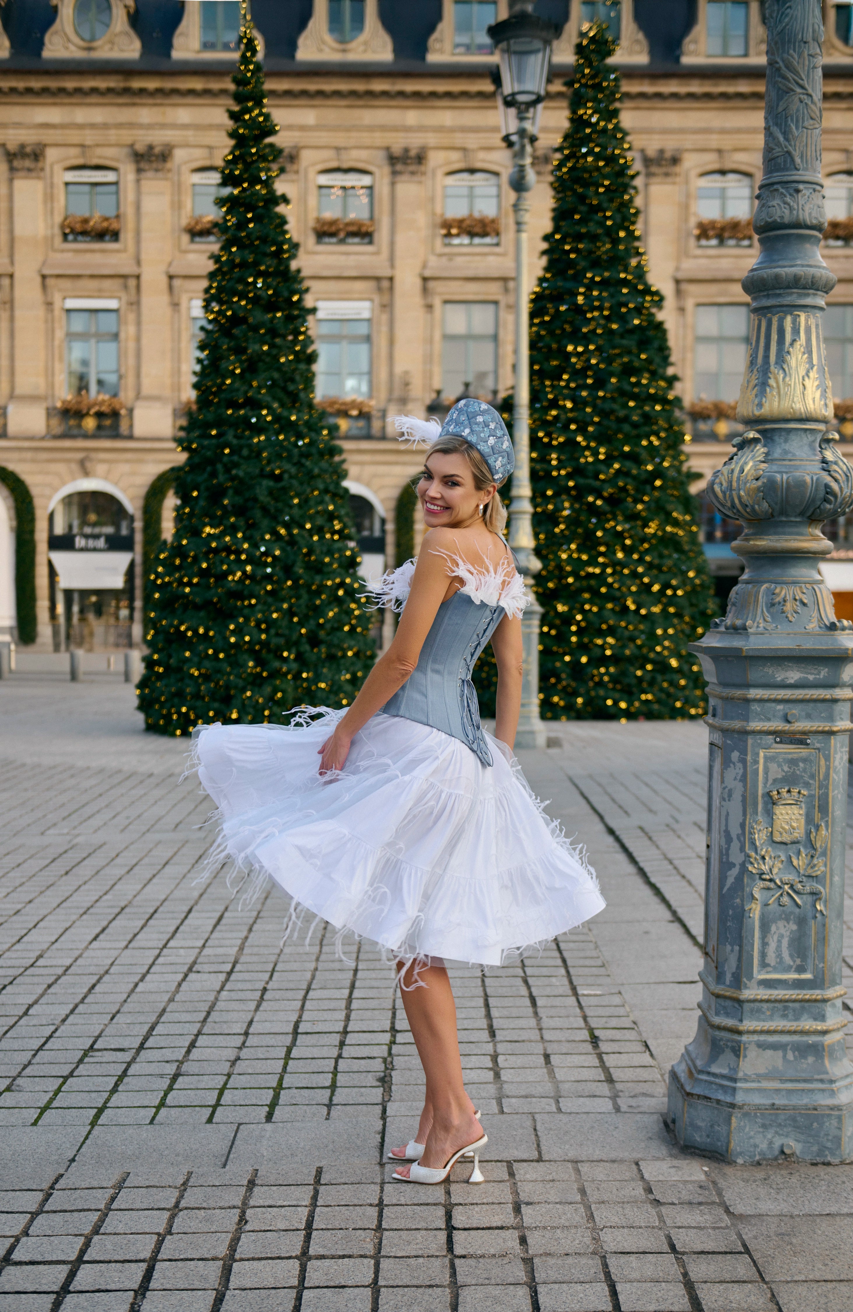 Woman in a light blue dress with white lace overlay standing on a paved square with Christmas trees in the background.