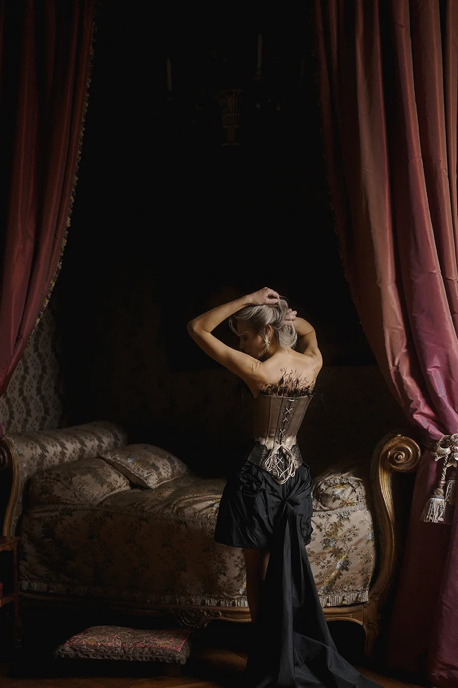 Woman in a gold corset by Mimaussy standing in a dimly lit room with ornate furniture and draped curtains.
