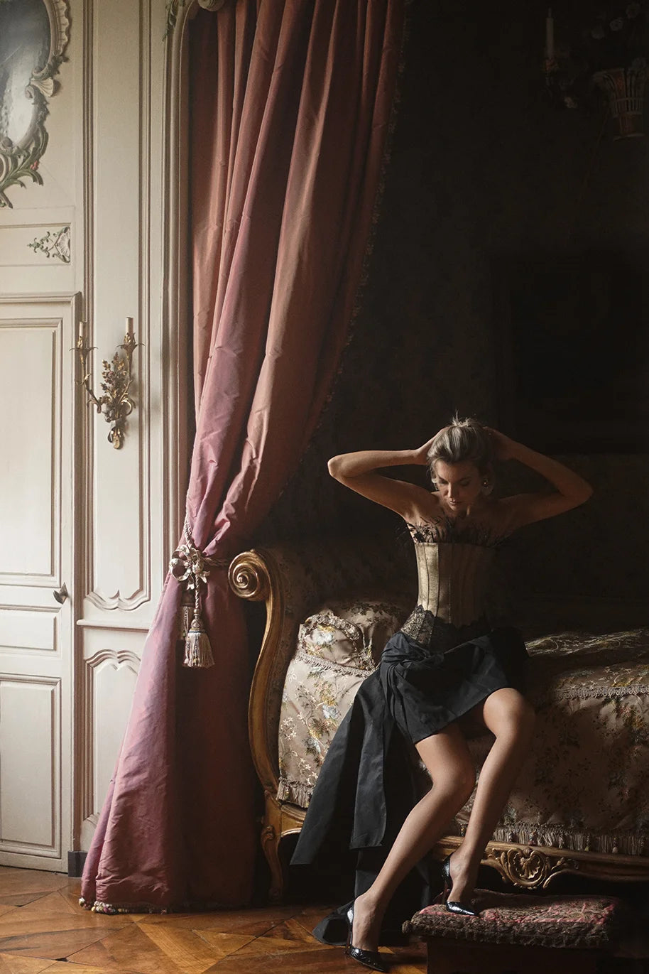 Woman wearing Gold Corset from Mimaussy Paris sitting on an ornate couch in a room with pink curtains and decorative wall elements.