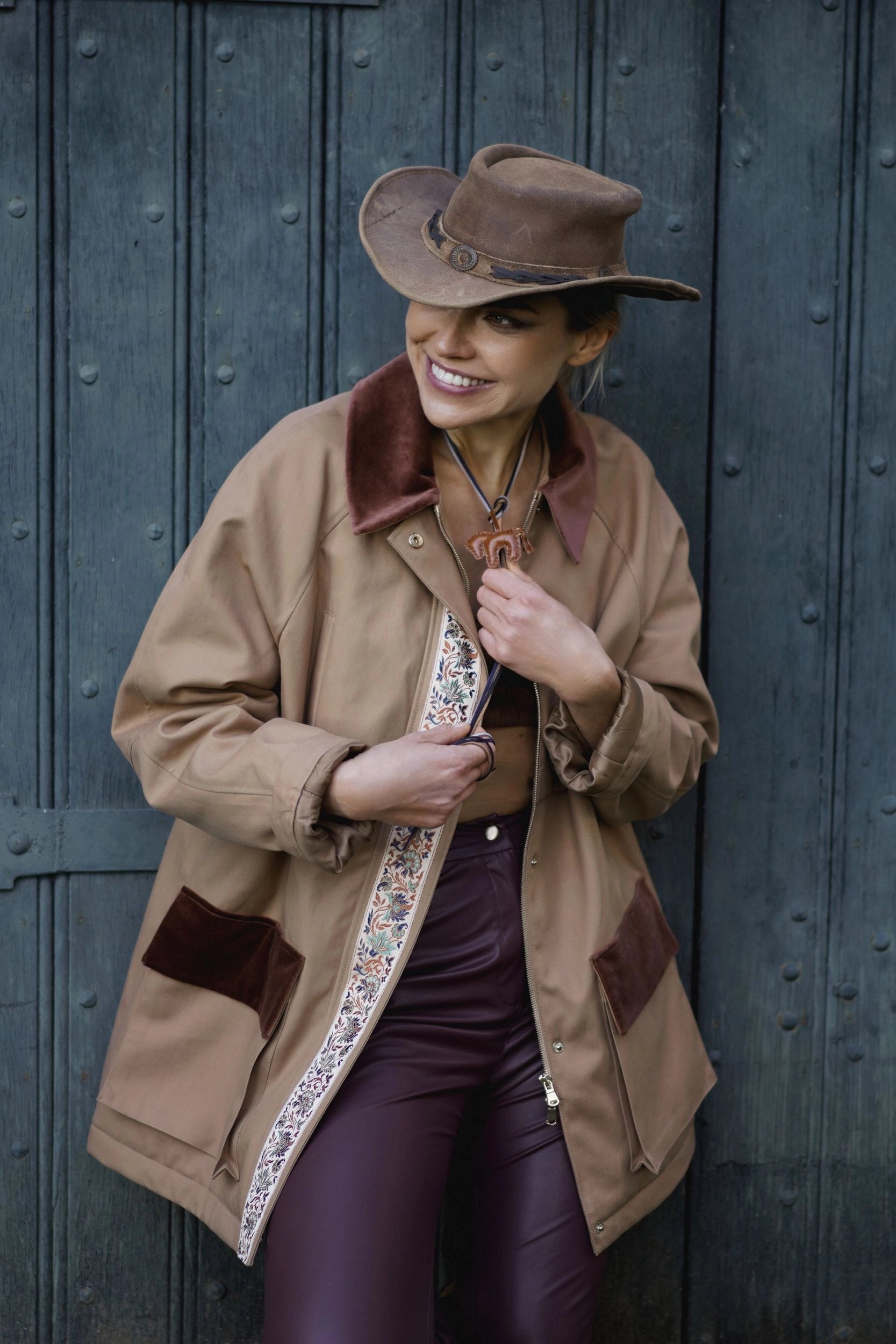 Woman in a tan coat and cowboy hat standing against a dark wooden door.