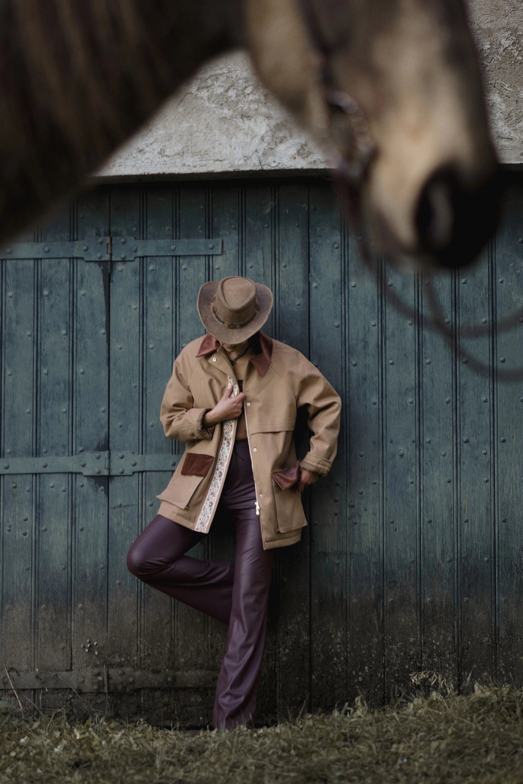 Person in vintage clothing leaning against a wooden shed with a horse in the foreground.