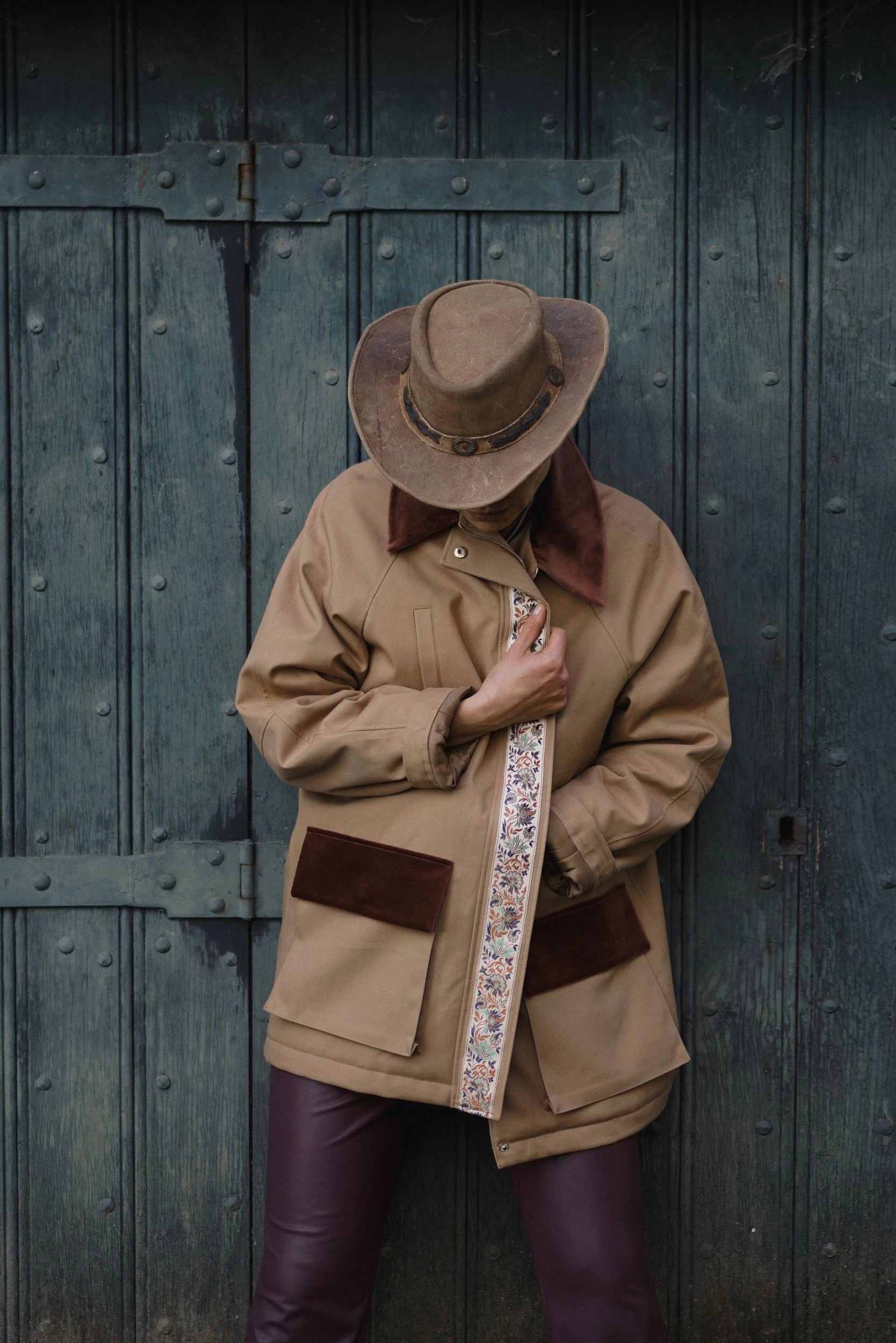 Person wearing a beige coat with brown accents and a hat, standing against a dark wooden door.