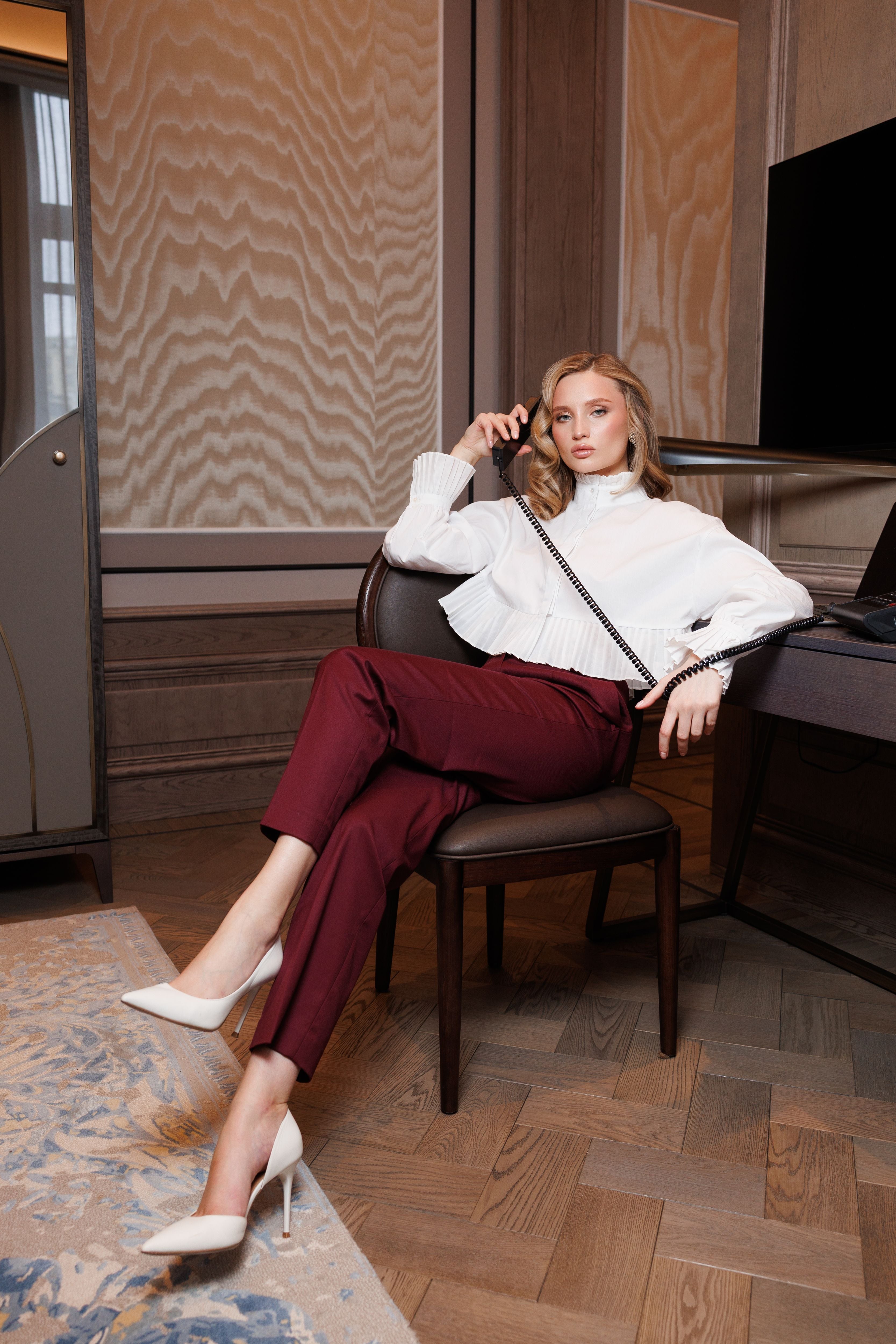 Woman in a white blouse and red pants sitting on a chair in a room with wooden flooring and a television.