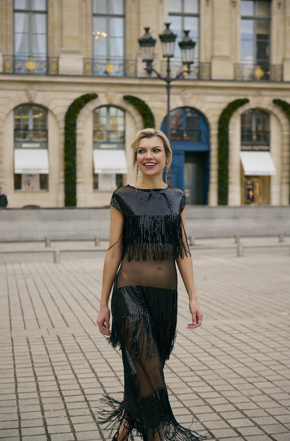 A woman in a black fringe dress walks confidently down the street  with classical architecture in the background, showcasing the Midnight Charleston Dress from Mimaussy Paris.