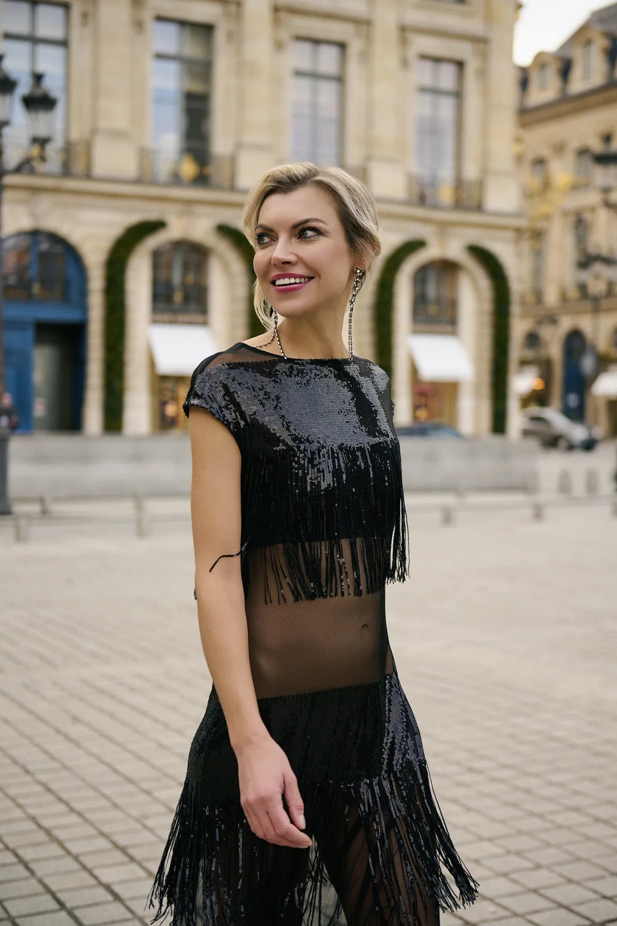 Close-up of a woman in a black fringe dress walks confidently down the street  with classical architecture in the background, showcasing the Midnight Charleston Dress from Mimaussy Paris.
