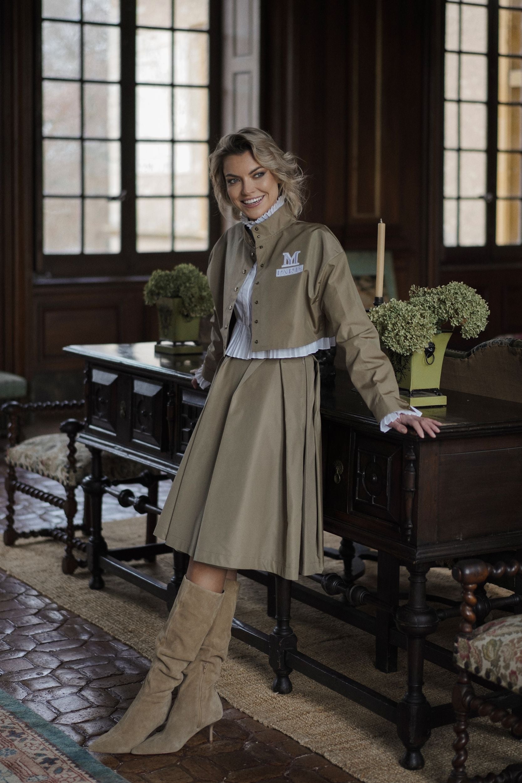 Woman wearing a Parisien Cropped Trench Jacket from Mimaussy Paris, beige jacket and skirt, standing in an elegant room with wooden furniture and large windows