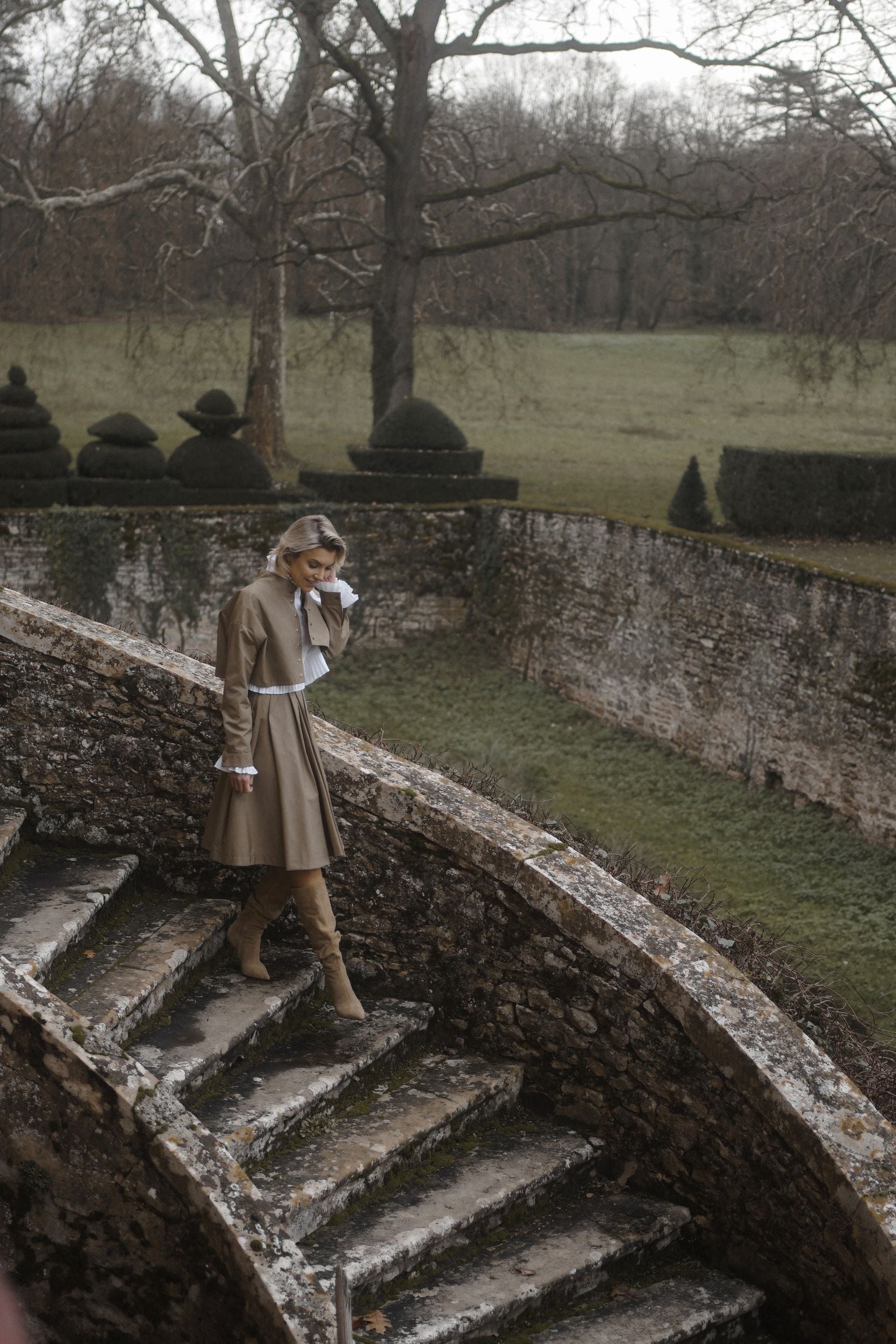 Woman wearing a Parisien Cropped Trench Jacket from Mimaussy Paris, long beige coat, standing on stone steps in a garden setting