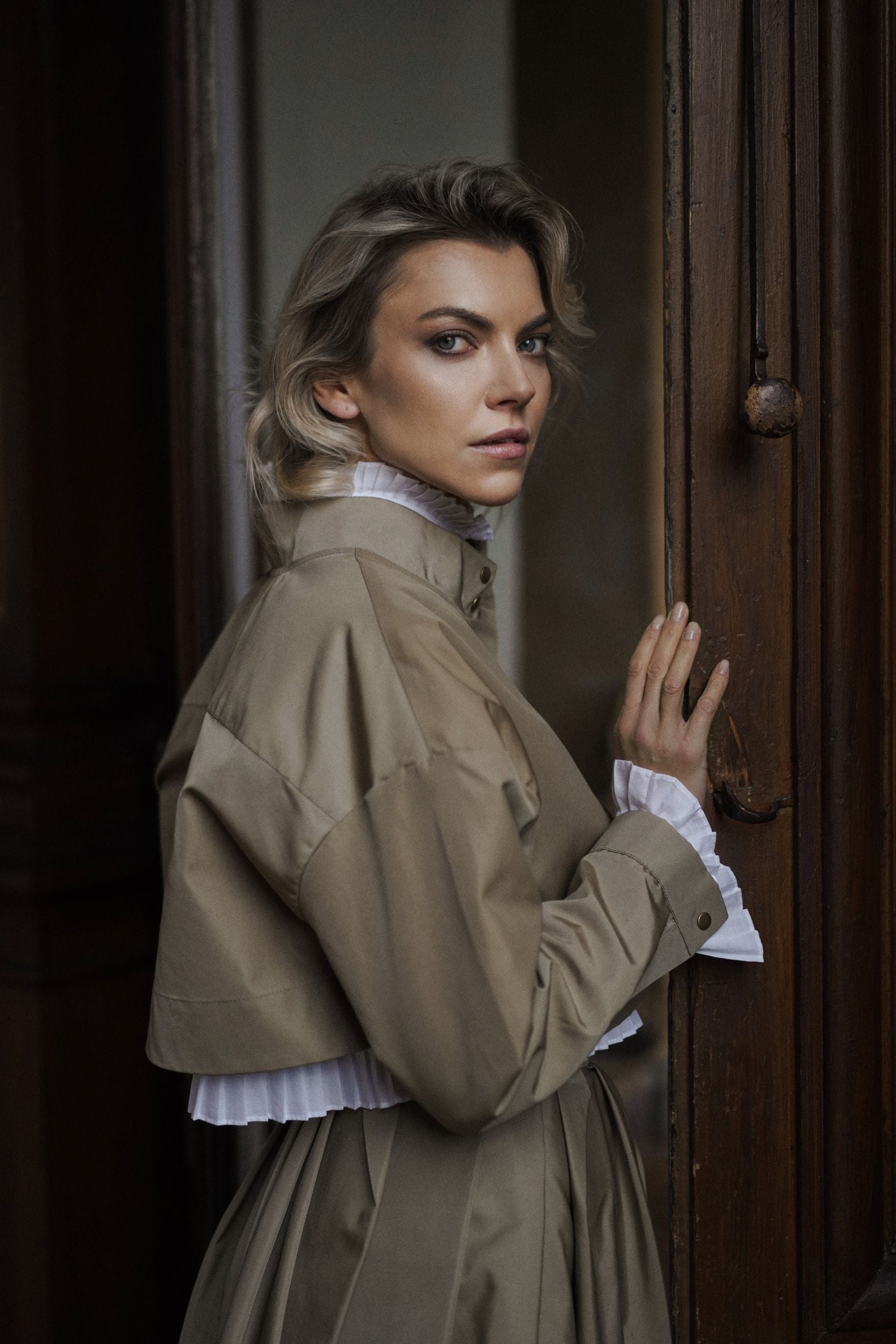 Woman wearing a Parisien Cropped Trench Jacket from Mimaussy Paris, beige coat, standing in a doorway with a dark background