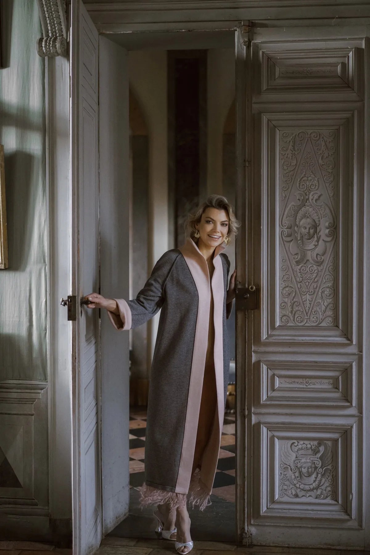 A woman in a Robe Coat with Ostrich Feather from Mimaussy Paris stands elegantly in an open doorway.
