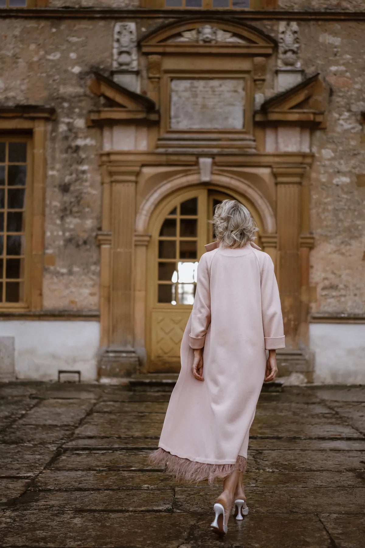 A woman in a pink robe coat with ostrich feathers from Mimaussy walks in front of a building, showcasing a stylish urban look.