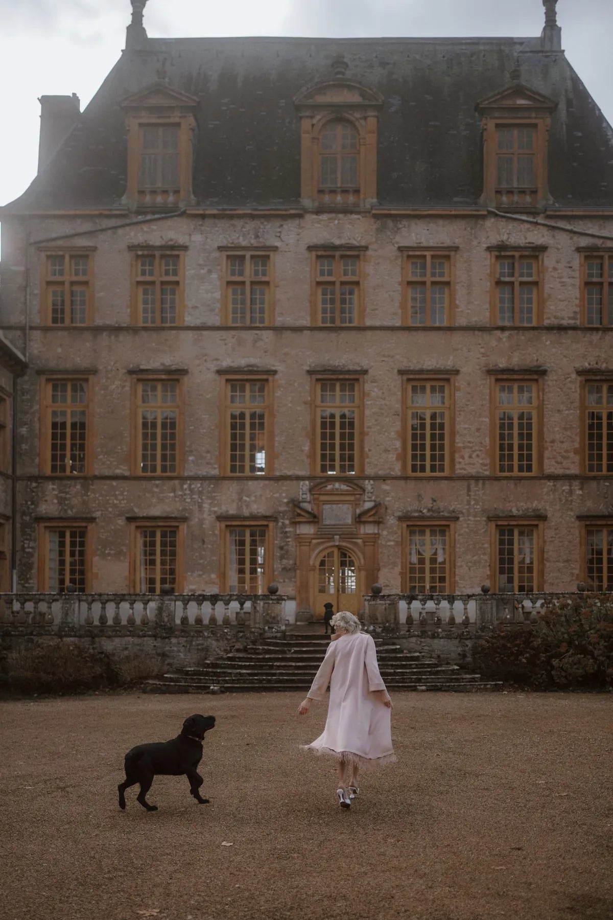 A model wearing the pink Robe Coat with Ostrich Feathers by Mimaussy Paris walks her dog in front of a large building.