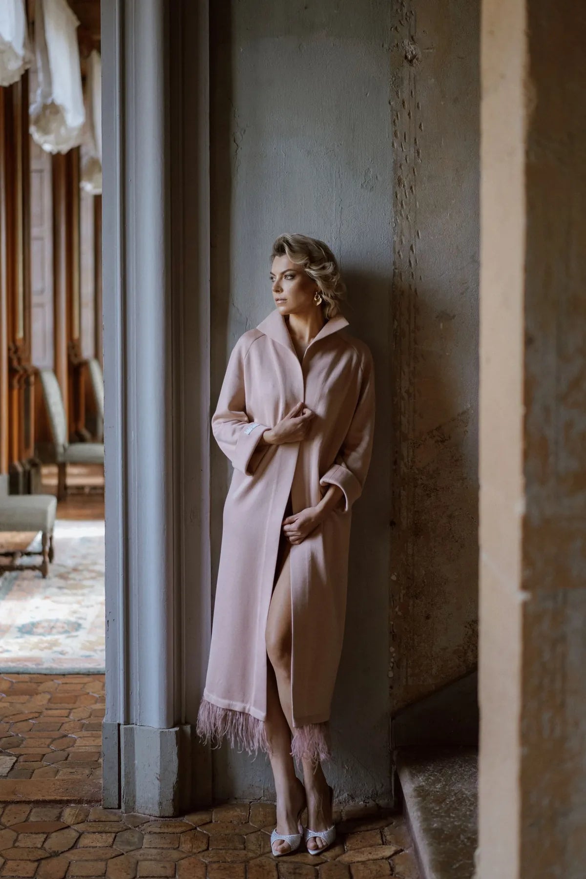 A woman wearing a pink Robe Coat with Ostrich Feathers from Mimaussy Paris leans against a wall in heels.