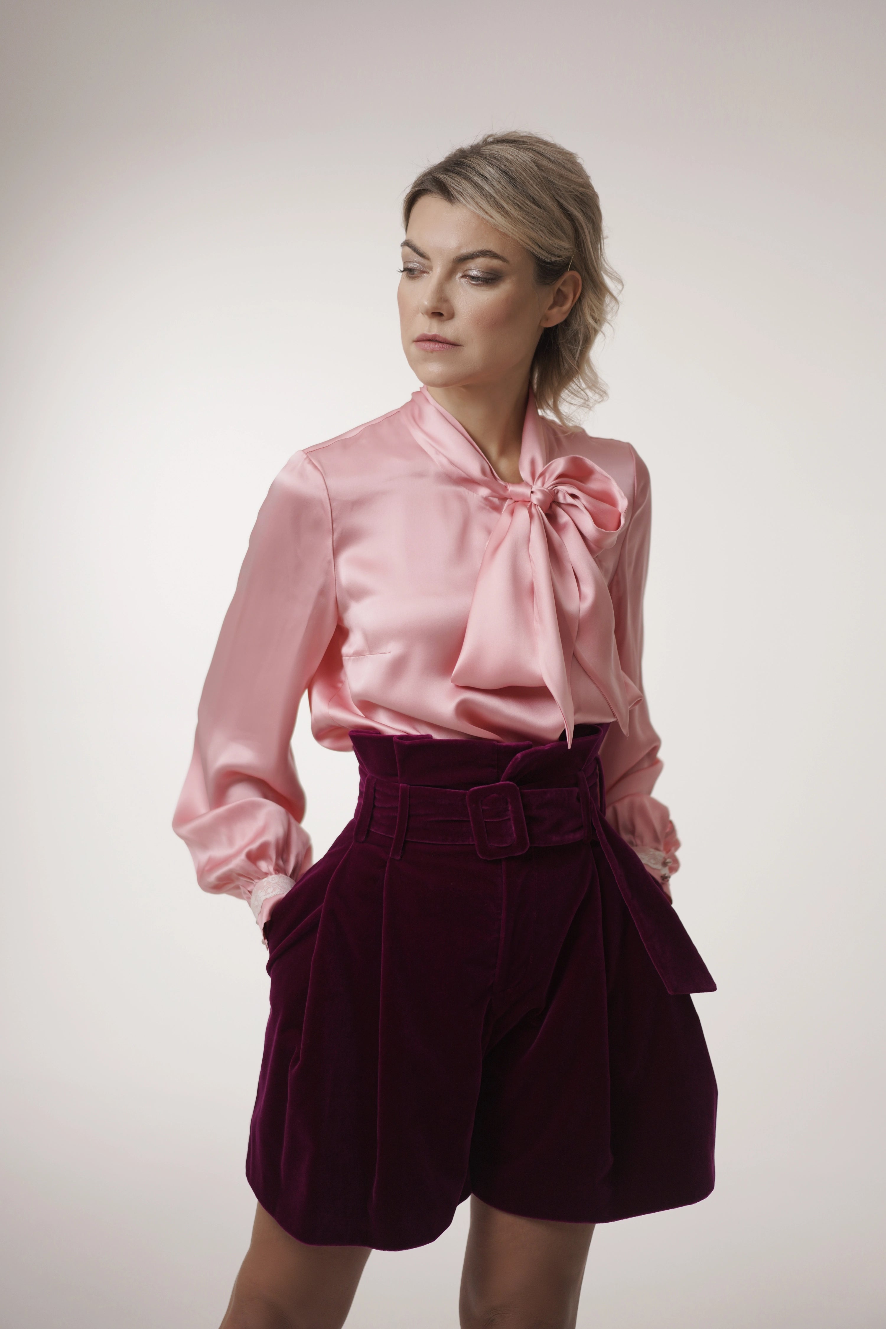 Woman wearing Rose Bow Blouse from Mimaussy Paris, pink blouse with bow paired with purple shorts on white background, studio setting