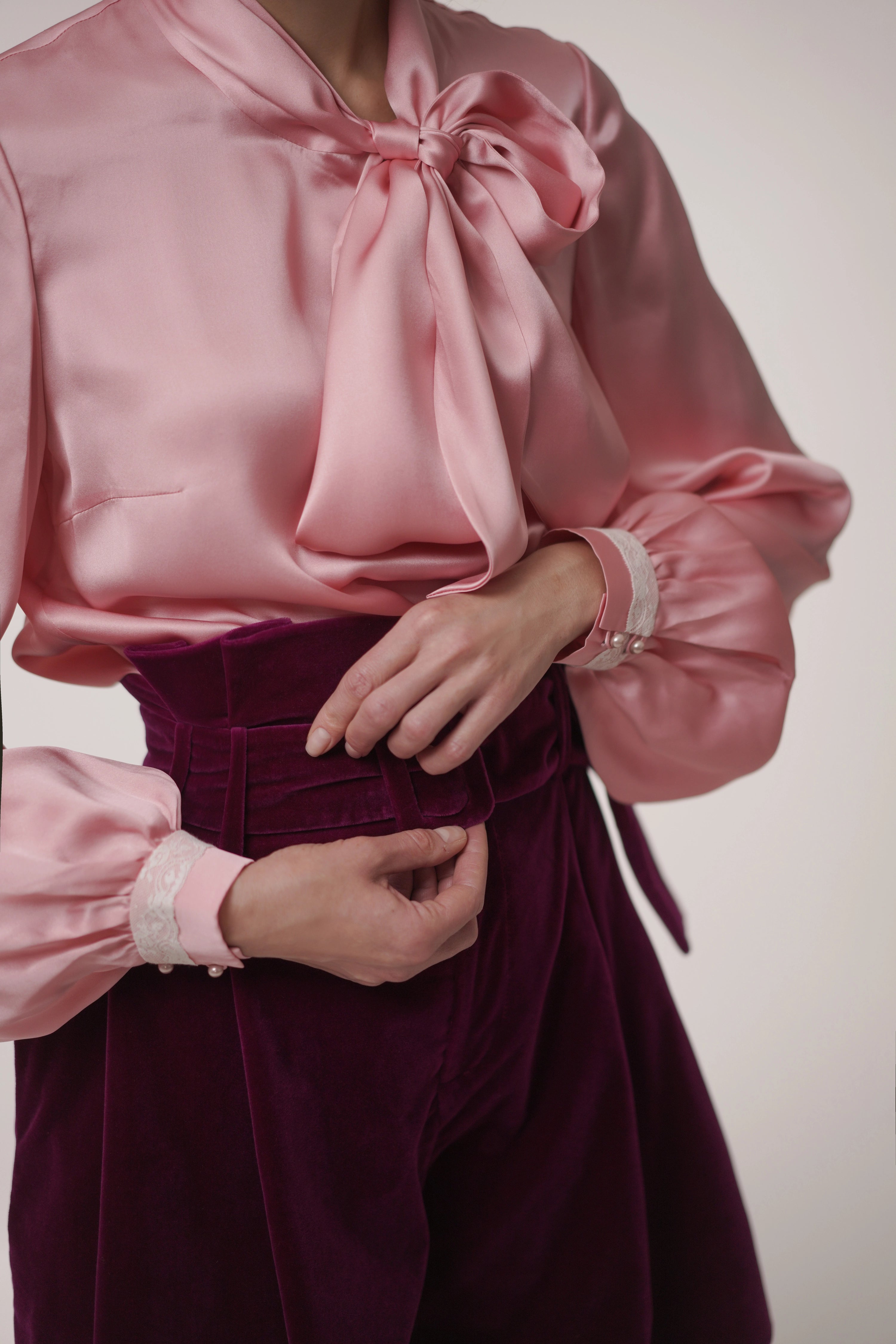 Woman wearing Rose Bow Blouse from Mimaussy Paris, pink satin blouse with large bow and burgundy pants on neutral background