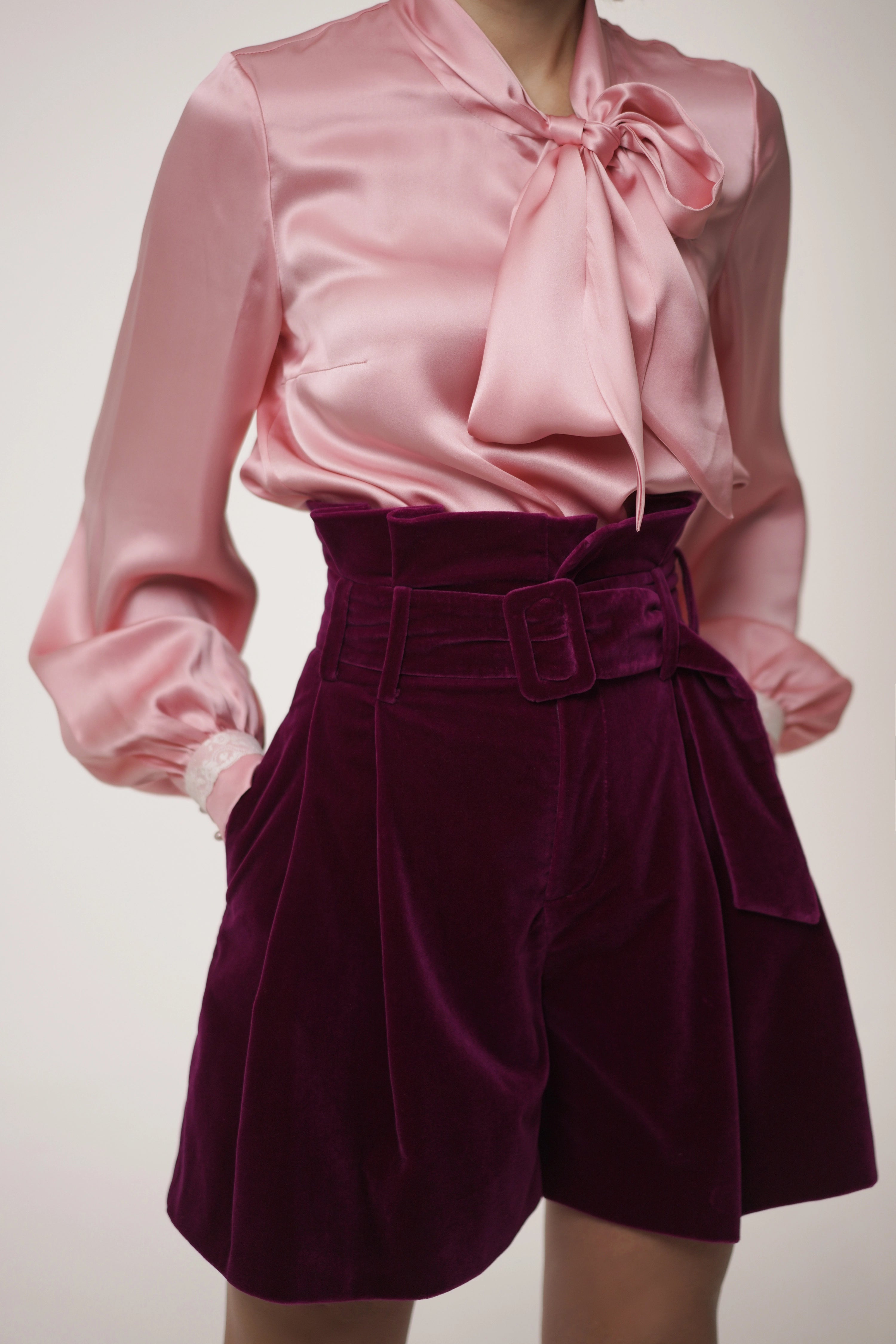 Woman wearing Rose Bow Blouse from Mimaussy Paris, pink satin blouse with large bow and purple velvet shorts on plain background