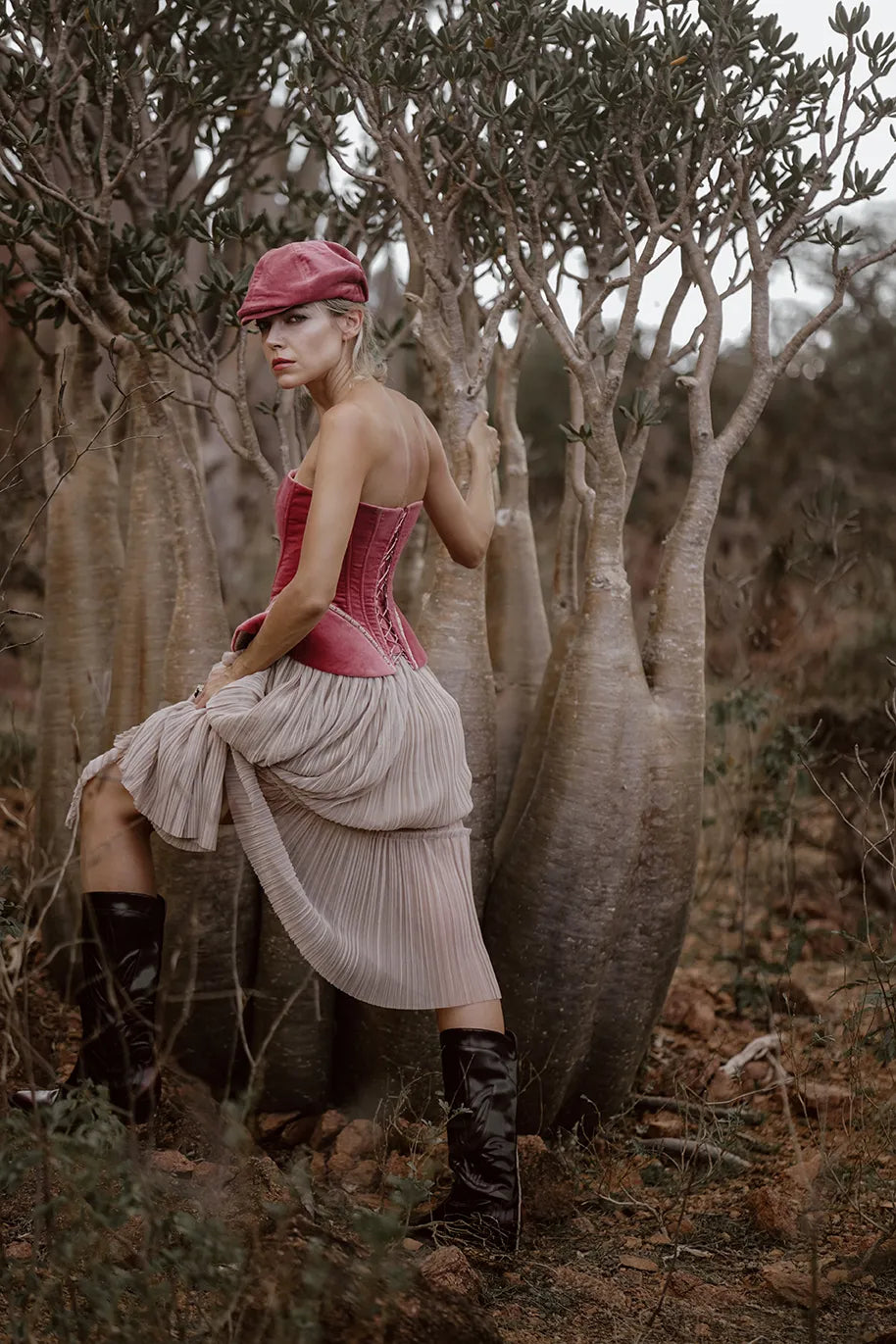 A woman in a pink hat and skirt poses gracefully in front of a tree, showcasing the luxury Sauvage Plissee Skirt from Mimaussy Paris.