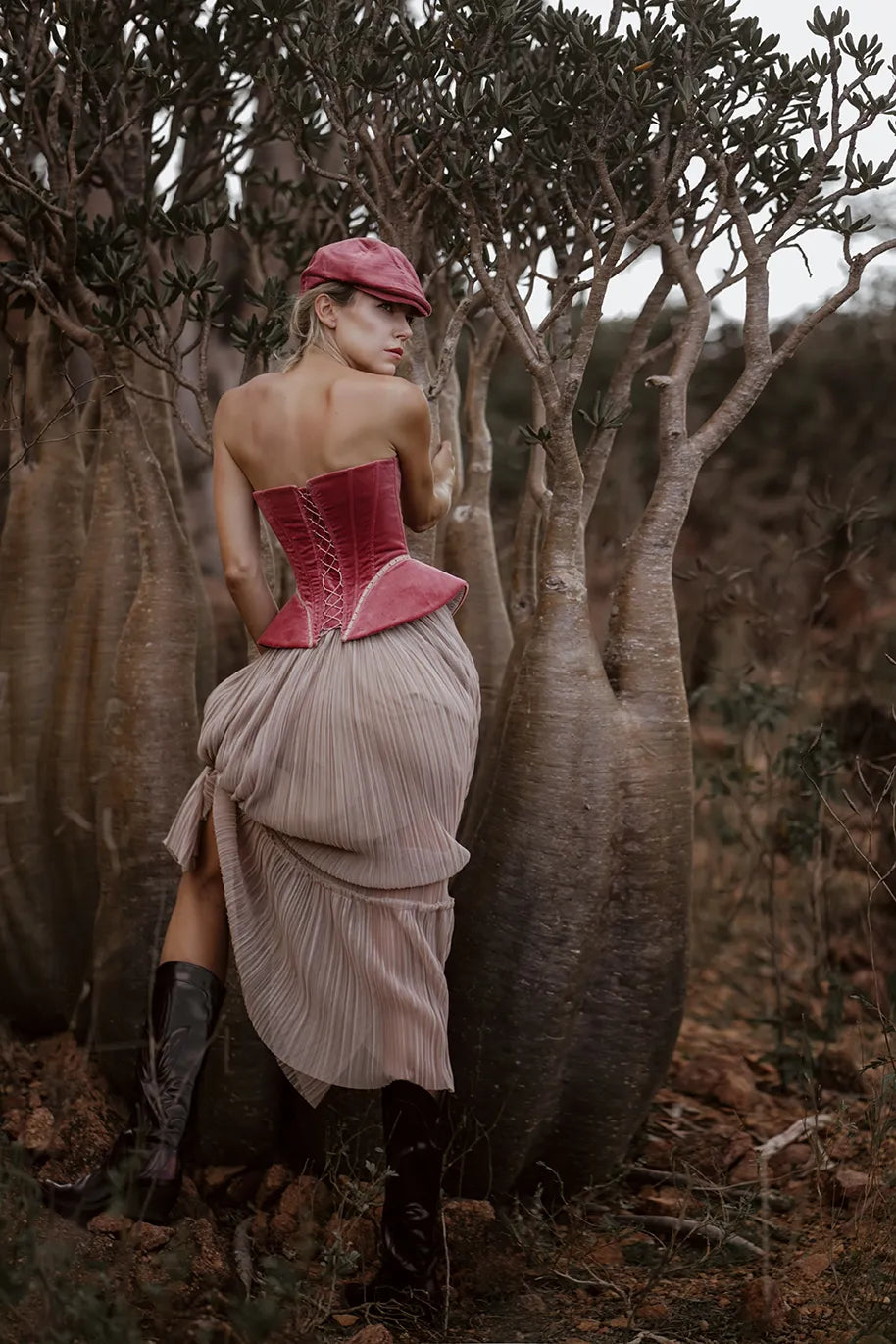 A woman in a Sauvage Plissee Skirt from Mimaussy Paris stands beside trees, wearing a stylish hat.
