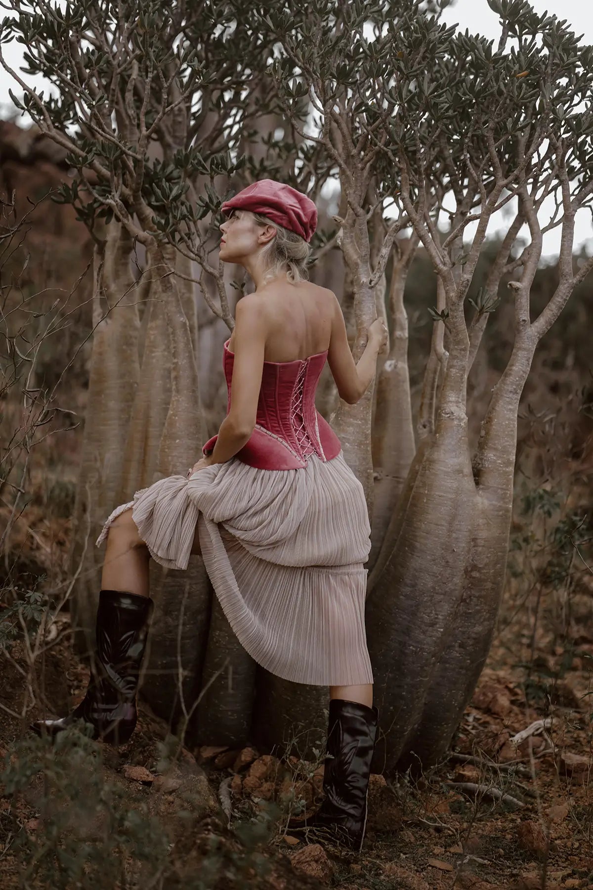 A woman in a luxury Sauvage Plissee Skirt from Mimaussy Paris stands beside a tree, wearing a stylish hat.