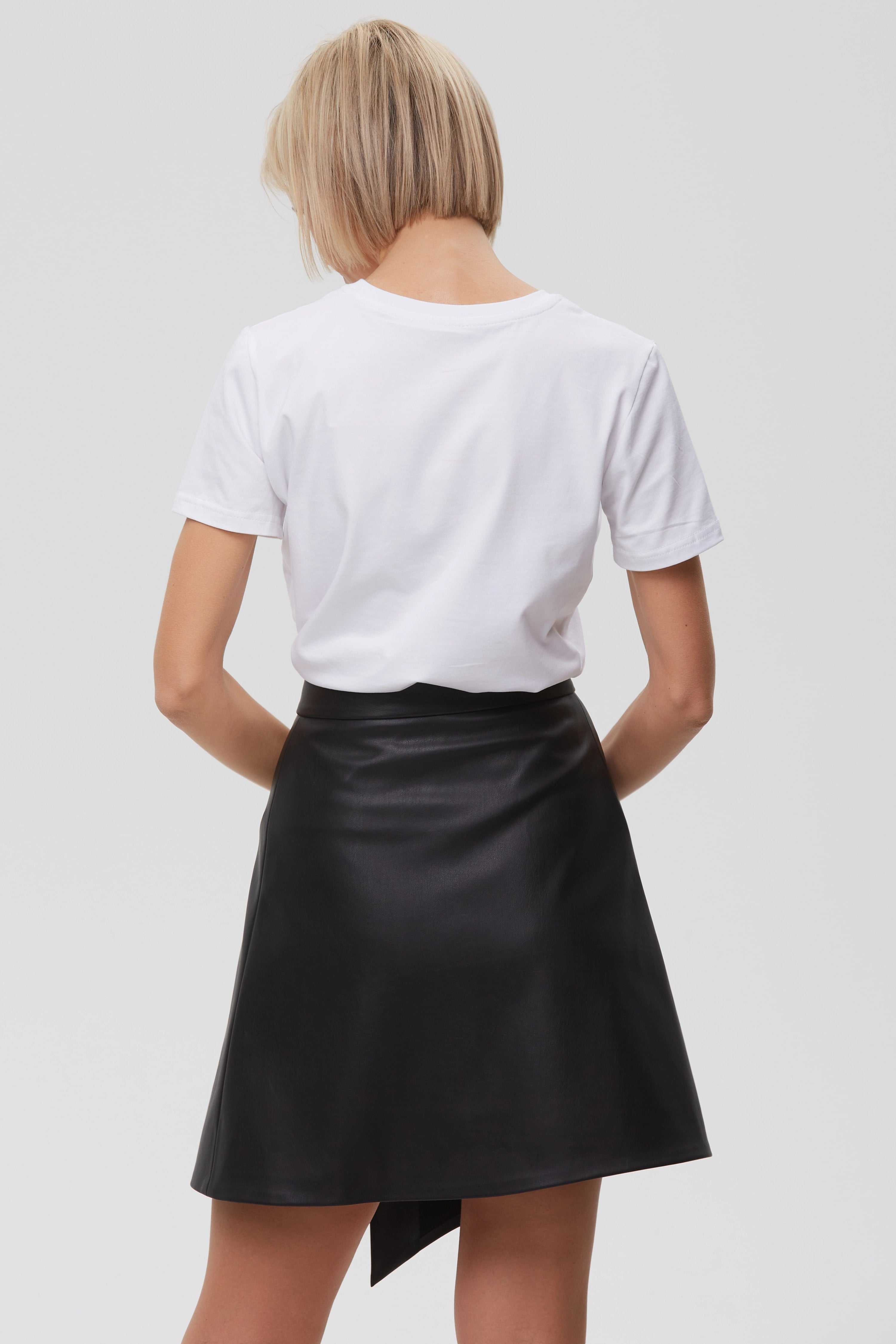 Woman wearing Skirt “Ligne” from Mimaussy Paris, back view of black leather skirt with white t-shirt on plain background