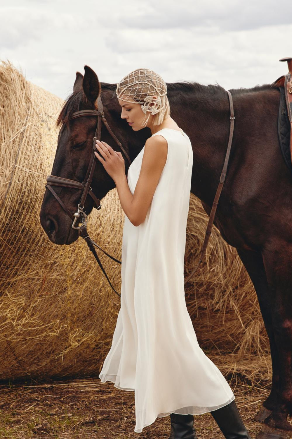 Woman in a white dress standing next to a horse in a rustic setting with hay bales.