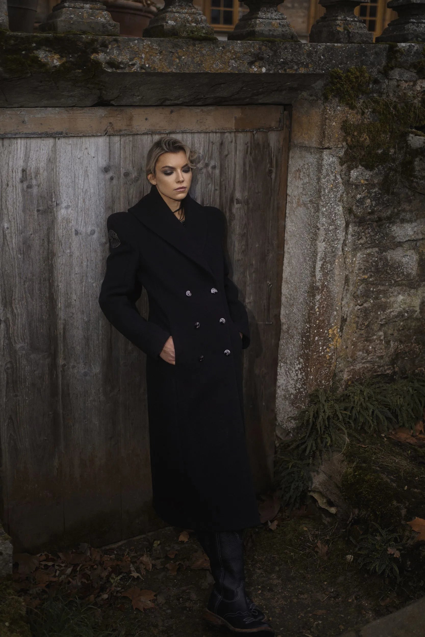 A woman wearing a black coat from Mimaussy Paris and boots stands before a wooden door, exuding elegance