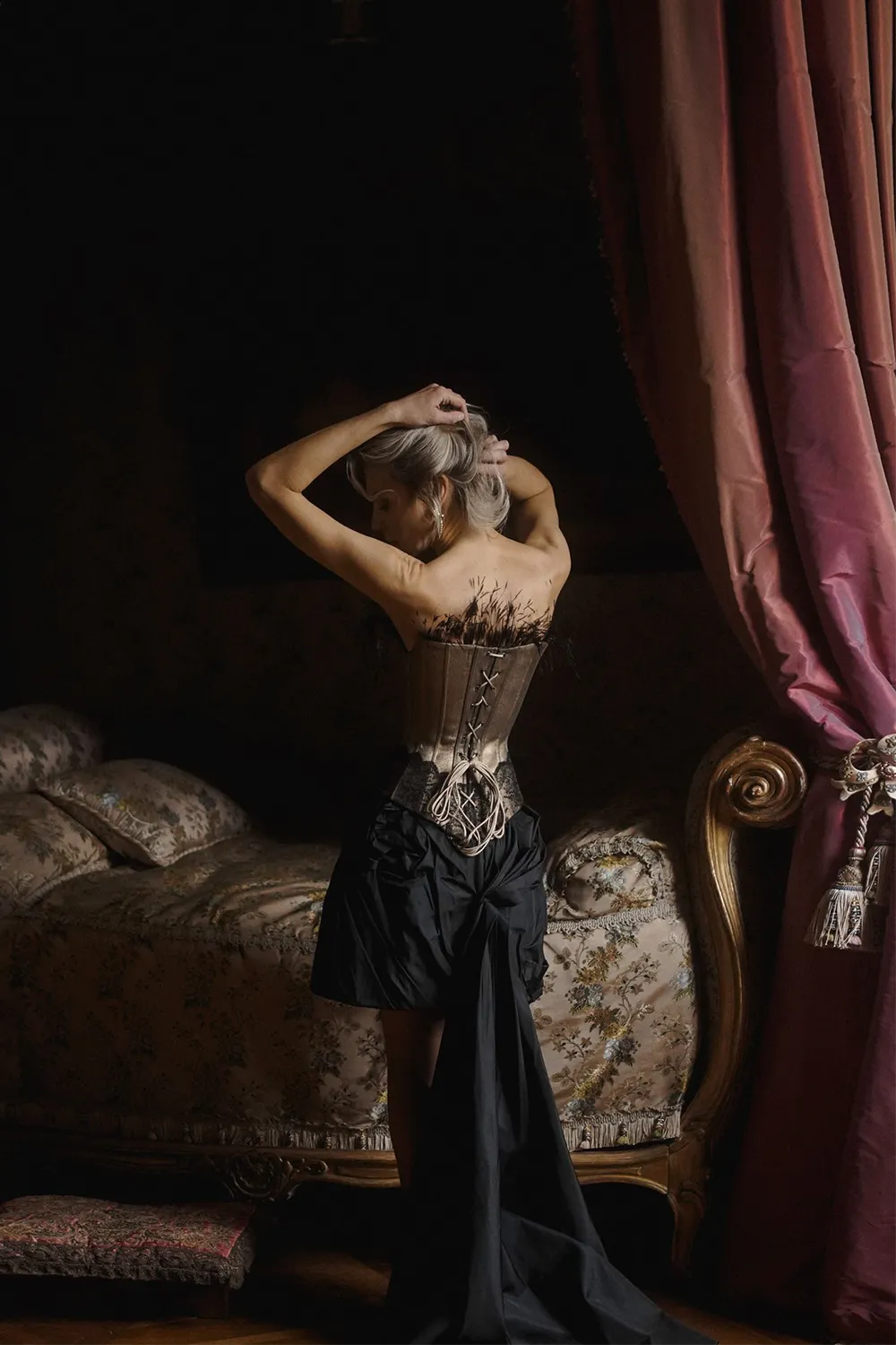 Lady in a Gold denim black corset and skirt standing in a dimly lit room with a bed and curtains.